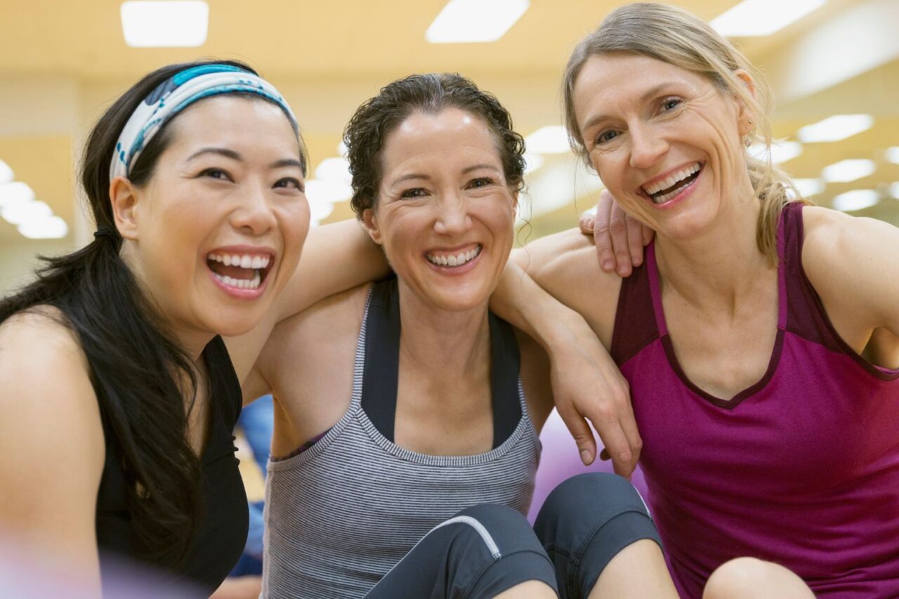 Three women in a group fitness class smiling at the camera at a gym